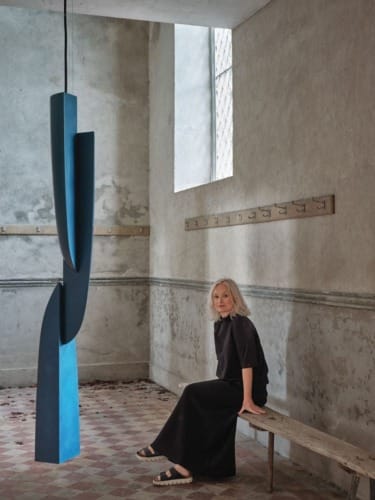 Woman seated on a wooden bench in a minimalist room with a large abstract blue sculpture hanging from the ceiling.