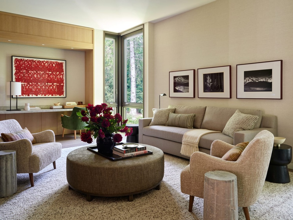 Modern living room with beige sofa, patterned chairs, round ottoman, and vibrant artwork on the wall. Large windows offer natural light.