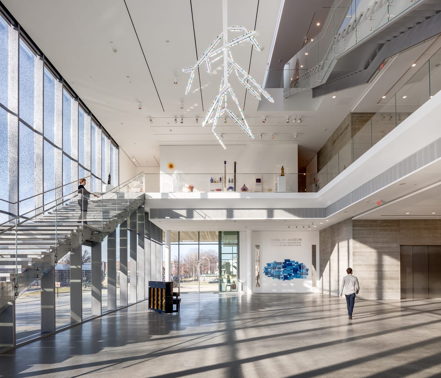 Modern art museum interior with large windows, staircase, hanging sculpture, and people walking on a sunny day.