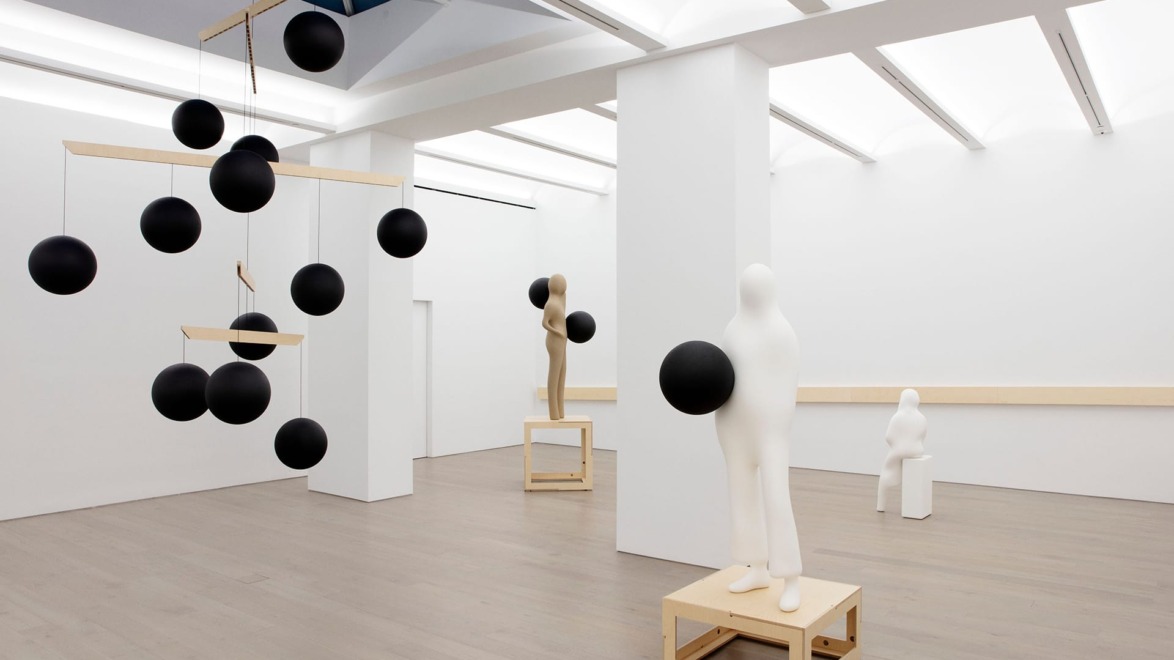 Minimalist art exhibit with abstract humanoid sculptures and black spherical elements in a brightly lit gallery space.