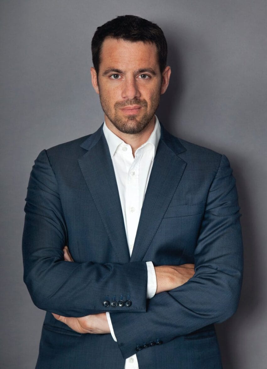 Man in a dark suit with arms crossed, standing against a gray background.