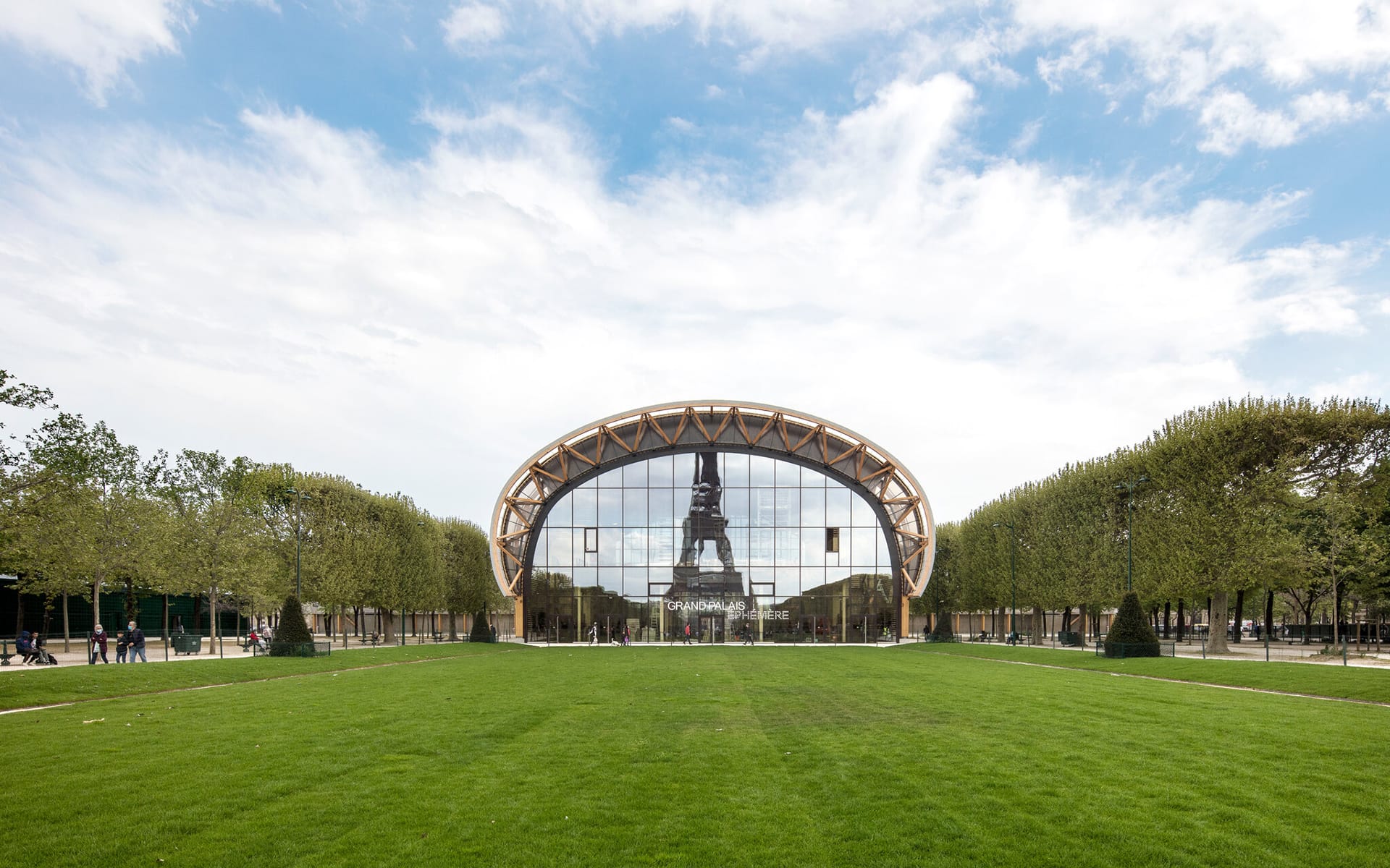 Modern building with glass facade reflecting part of the Eiffel Tower, surrounded by a lush green lawn and trees.