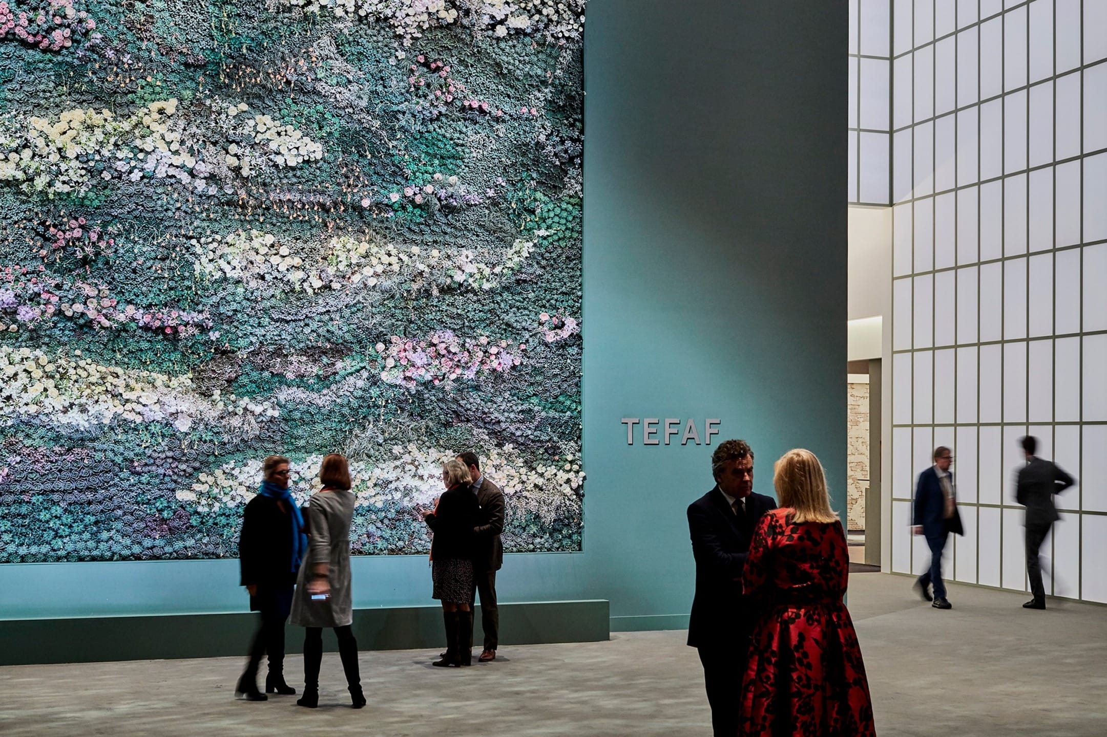 People interacting at an art fair with a large textured wall display in the background, labeled TEFAF, in a modern setting.