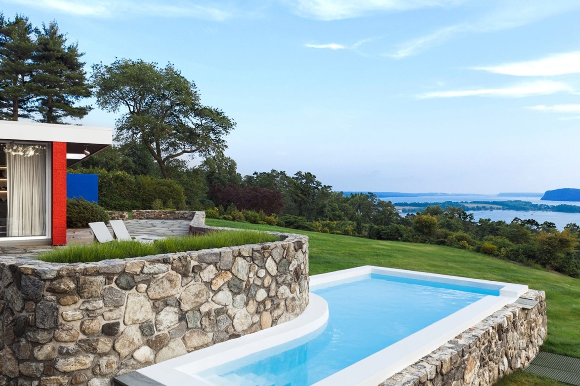 Modern hillside pool with stone wall, lush greenery, and lake view under a clear blue sky.
