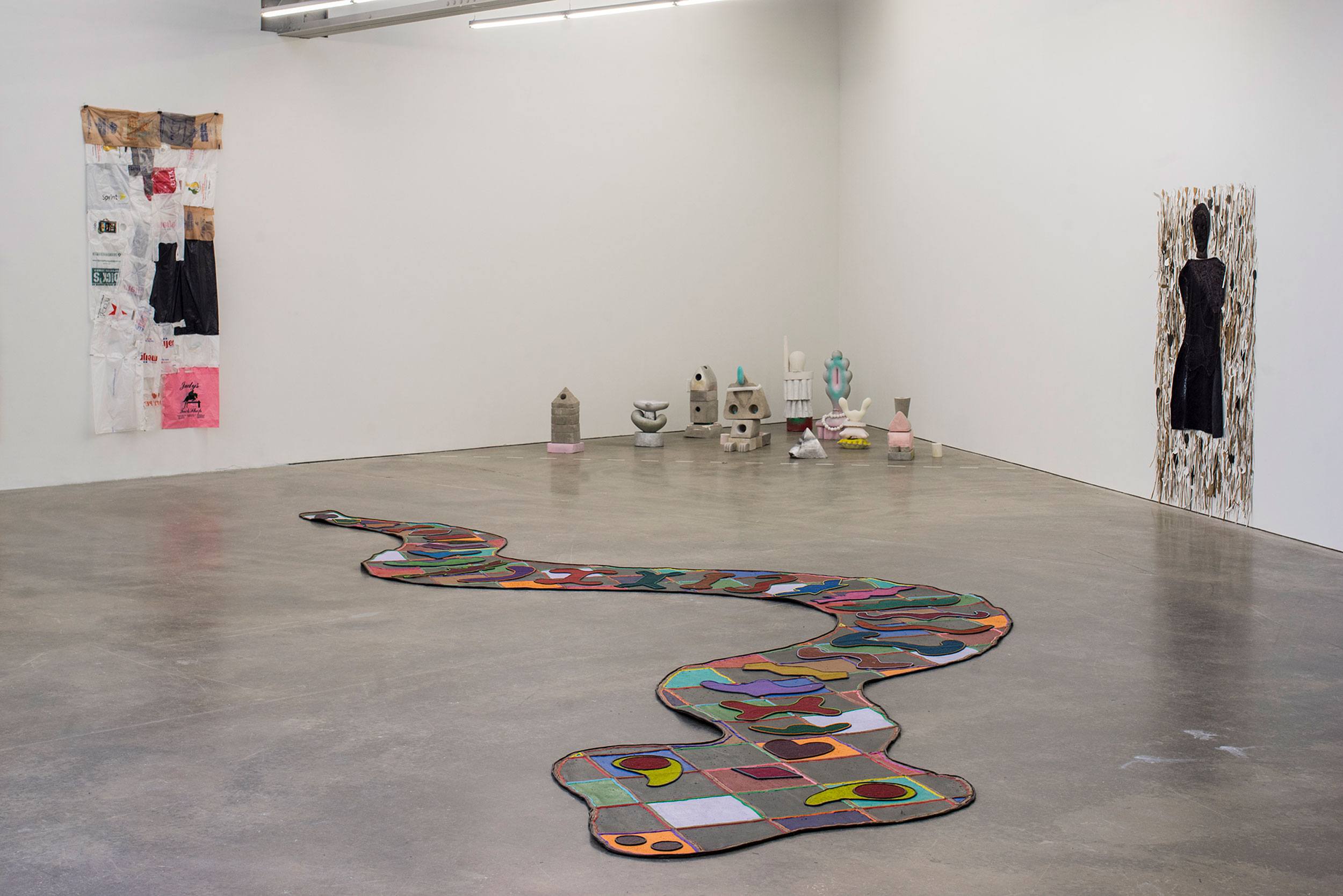 Art installation in a gallery with a patterned floor piece, sculptures, and wall hangings in a minimalist setting.
