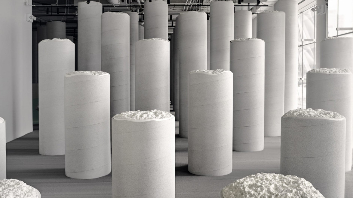 Modern sculpture installation with tall, cylindrical white pillars in an industrial-style gallery space with visible pipes above.