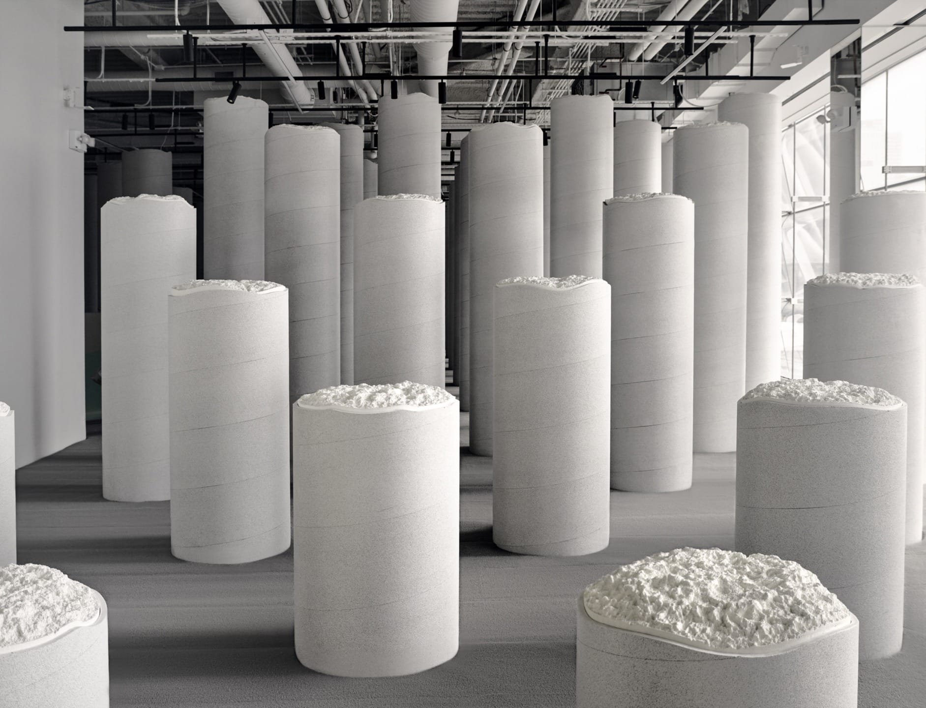 Modern sculpture installation with tall, cylindrical white pillars in an industrial-style gallery space with visible pipes above.