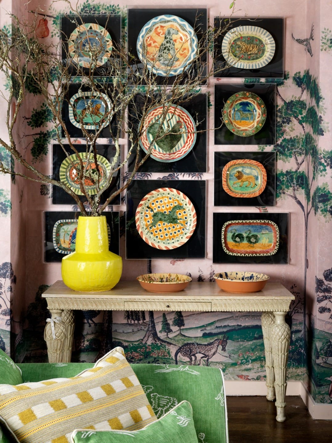 Colorful decorative plates on a wall above a wooden table with a bright yellow vase and green patterned cushions.