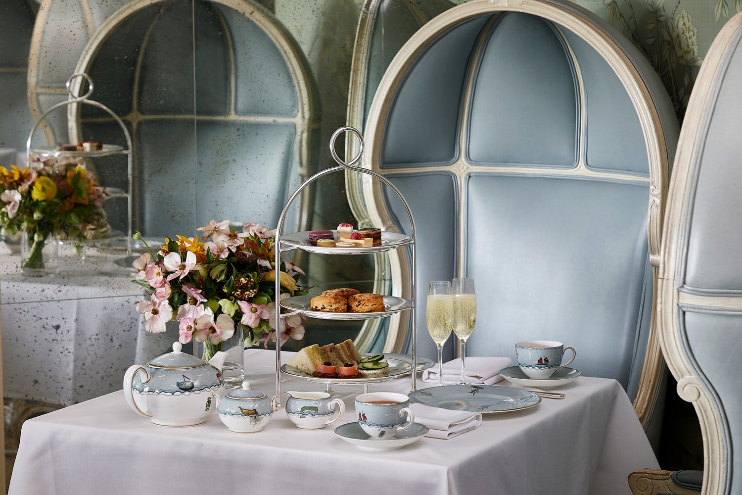 Elegant afternoon tea setup with pastries, tea set, and champagne glasses on a white tablecloth near floral arrangements.