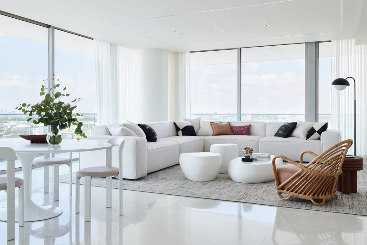 Modern living room with large windows, white sectional sofa, round coffee tables, wicker chair, and dining table.