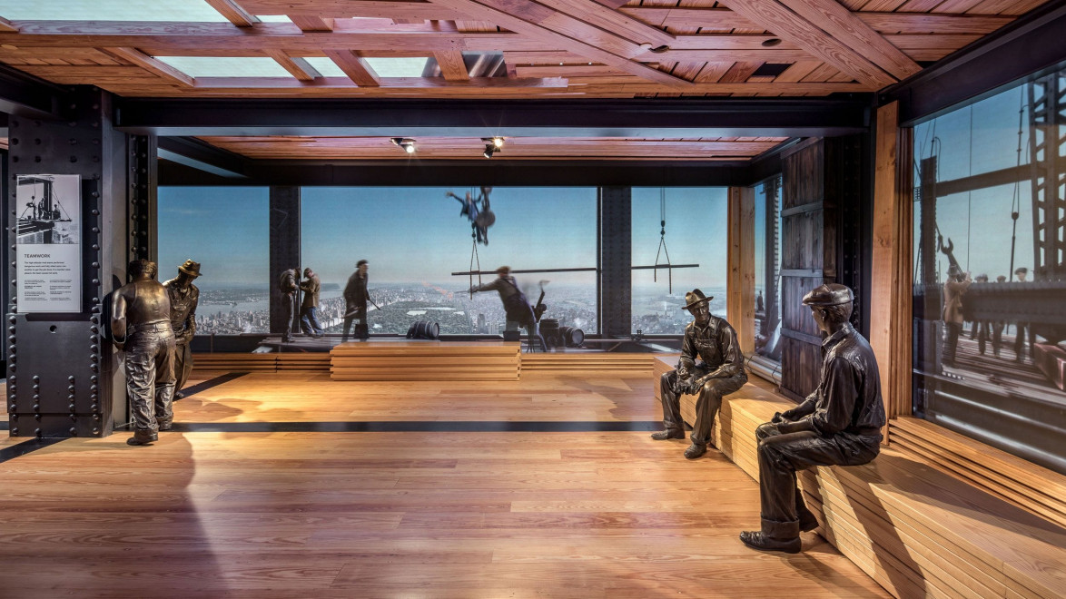 Museum exhibit with statues of construction workers sitting and standing on a wooden floor, large city skyline in background.