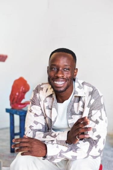 Smiling person sitting in a patterned jacket, with a red sculpture and blue stool in the background.