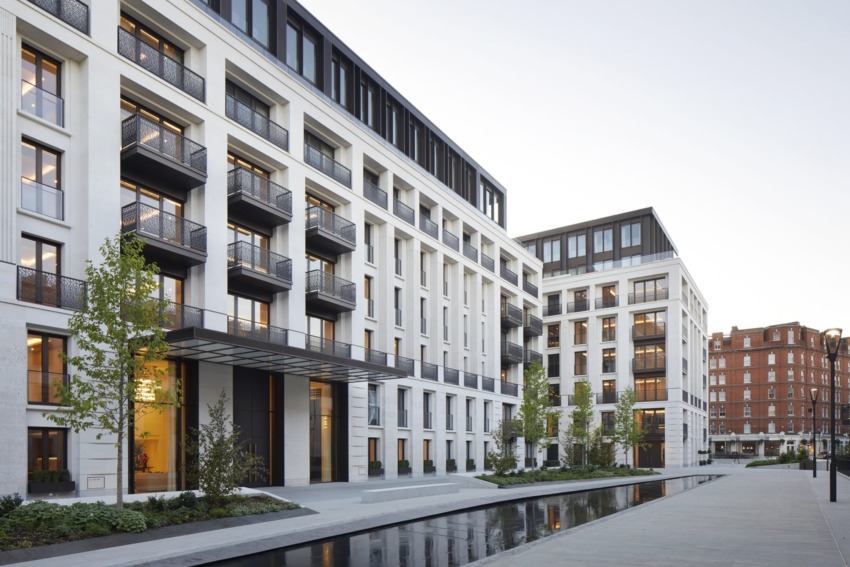Modern apartment complex with large windows, balconies, and a reflecting pool in an urban setting during daytime.
