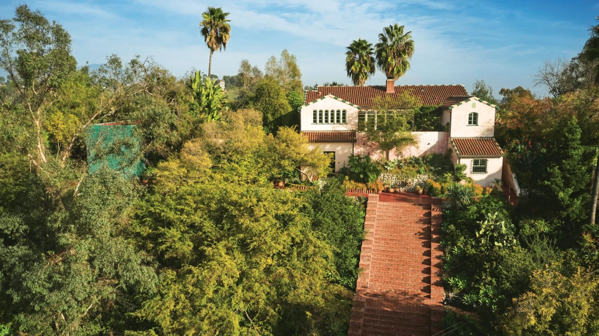 Spanish-style house surrounded by lush greenery and tall palm trees under a clear blue sky.