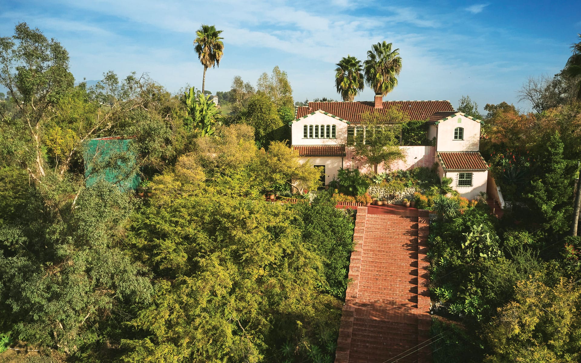 Spanish-style house surrounded by lush greenery and tall palm trees under a clear blue sky.