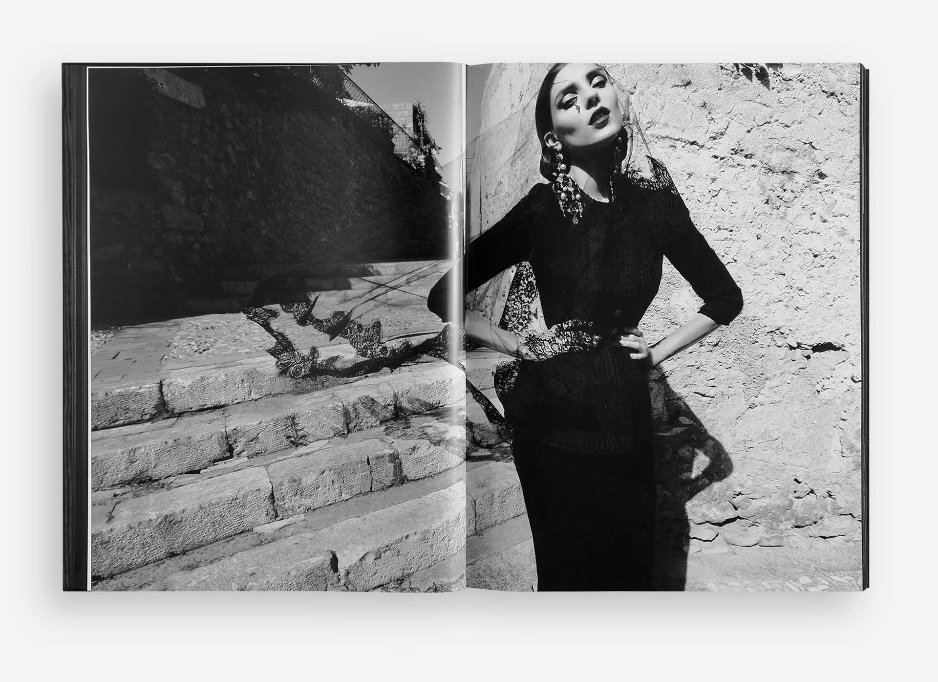 Black and white photo of a model posing in a stylish outfit near stone steps and a wall in an open fashion book.
