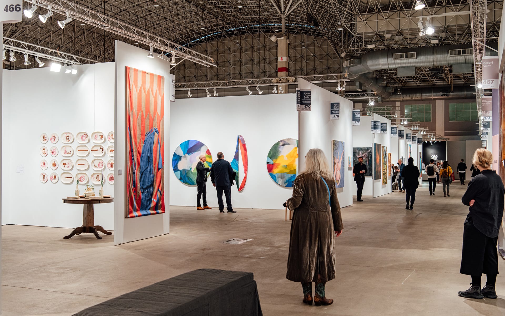 Art exhibition with people viewing colorful paintings and installations in a spacious gallery setting.
