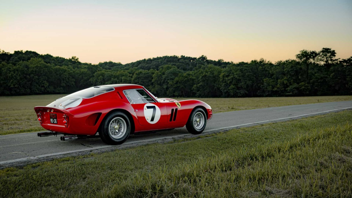 Classic red sports car with number 7 on the side, parked on a rural road with trees and a sunset in the background.
