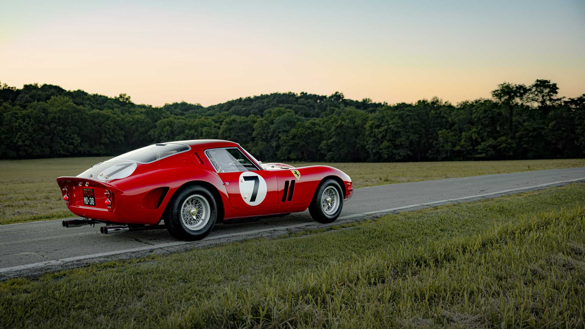 Classic red sports car with number 7 on the side, parked on a rural road with trees and a sunset in the background.