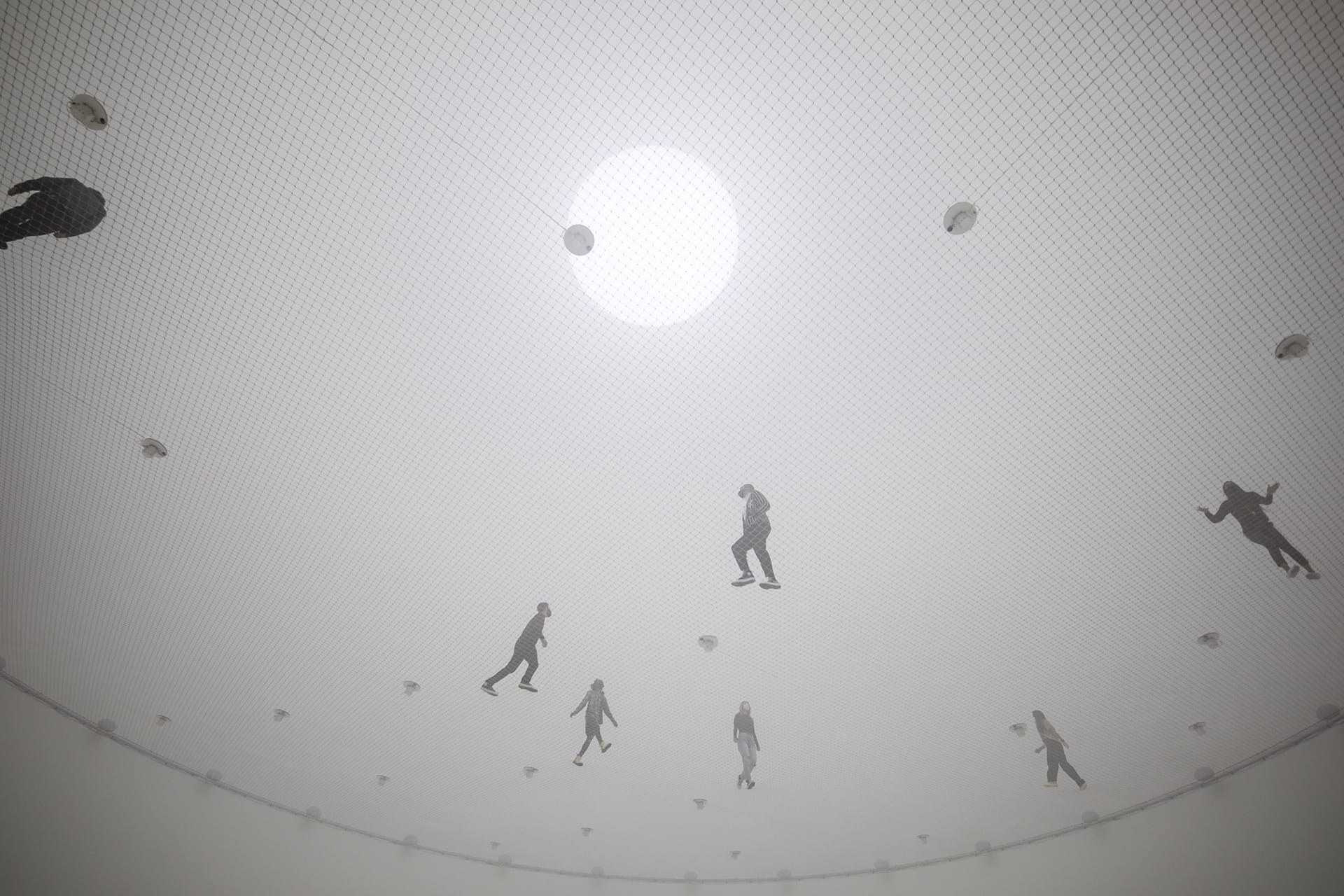 People walking on a net suspended in a foggy environment with the sun visible through the mist above them.