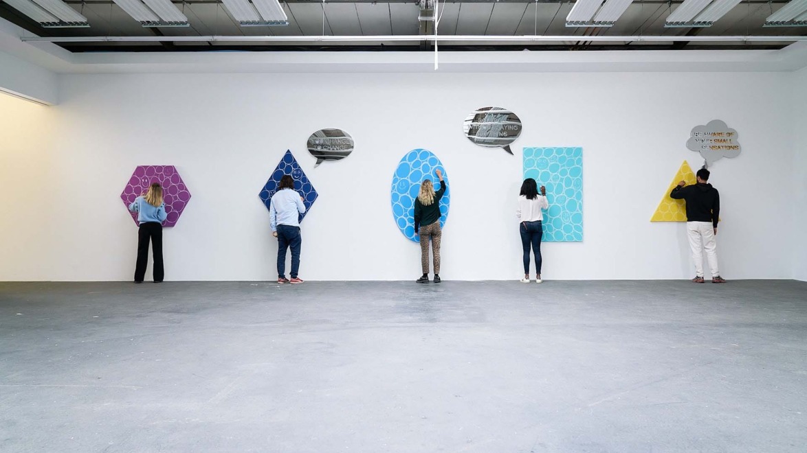 People interacting with geometric art pieces mounted on a gallery wall under bright ceiling lights.
