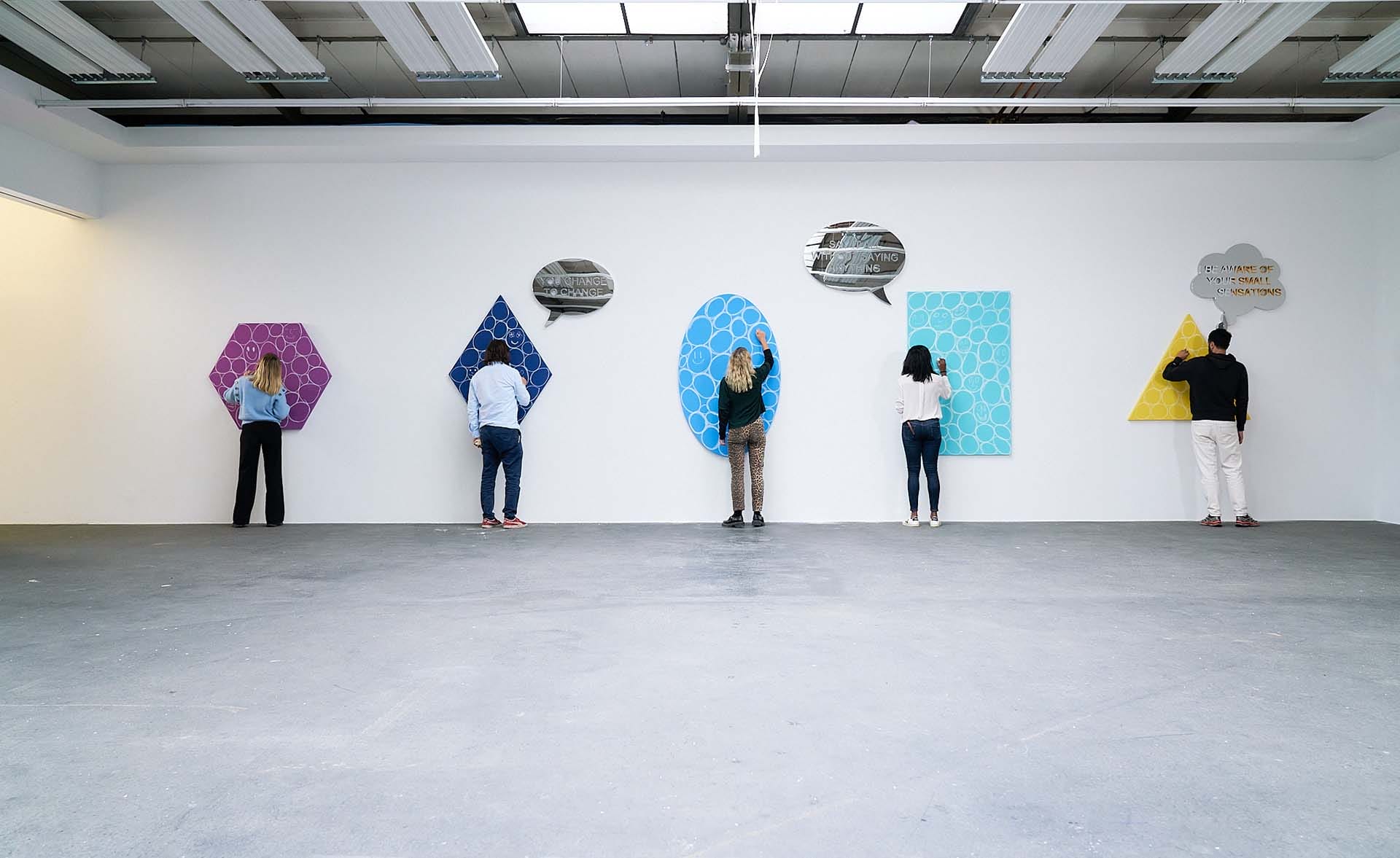 People interacting with geometric art pieces mounted on a gallery wall under bright ceiling lights.