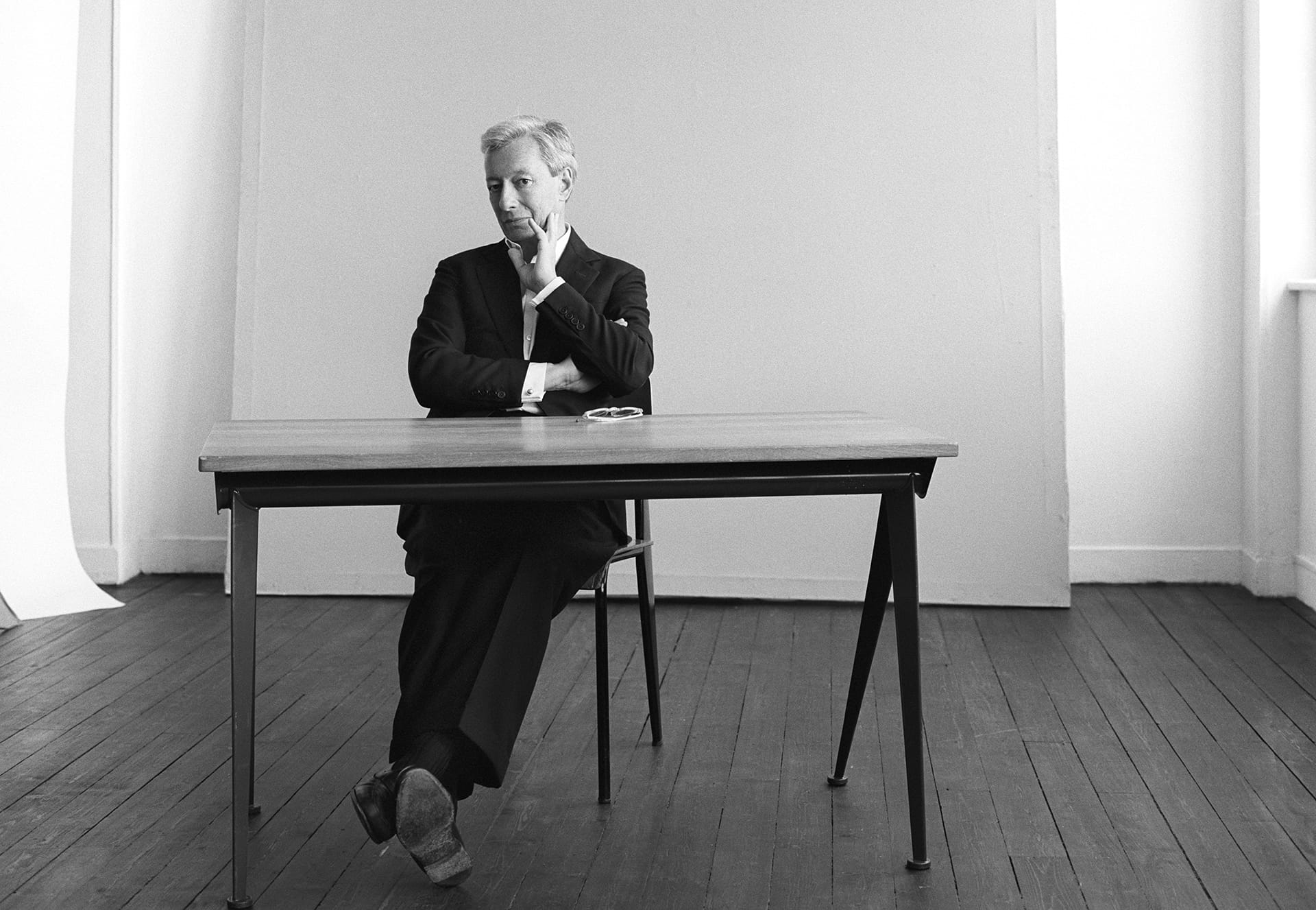 Man sitting thoughtfully at a wooden table in a minimalist studio setting, with a neutral backdrop and hardwood floor.