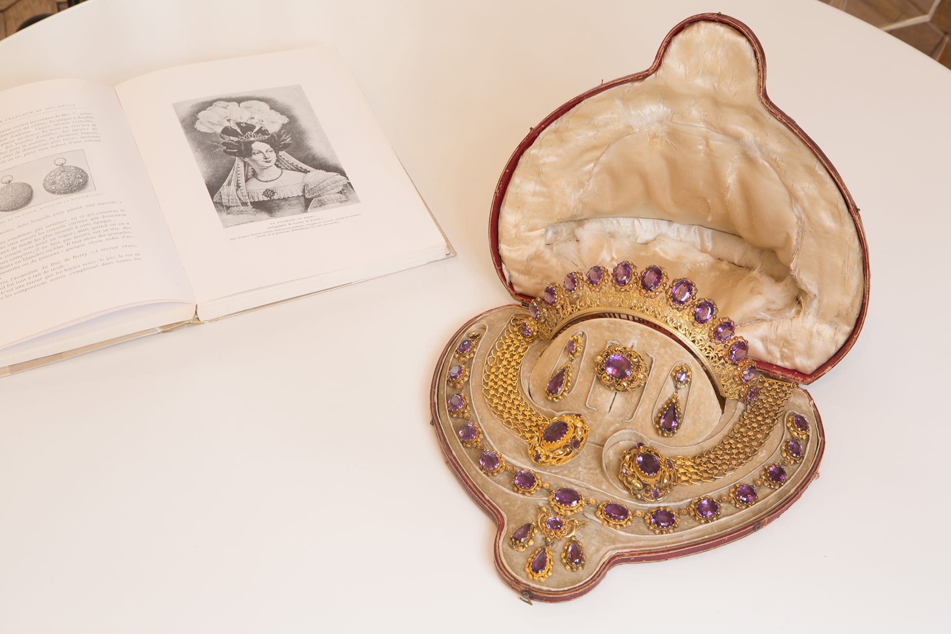 Vintage jewelry set with purple gems in an ornate case next to an open book showing a historical illustration.