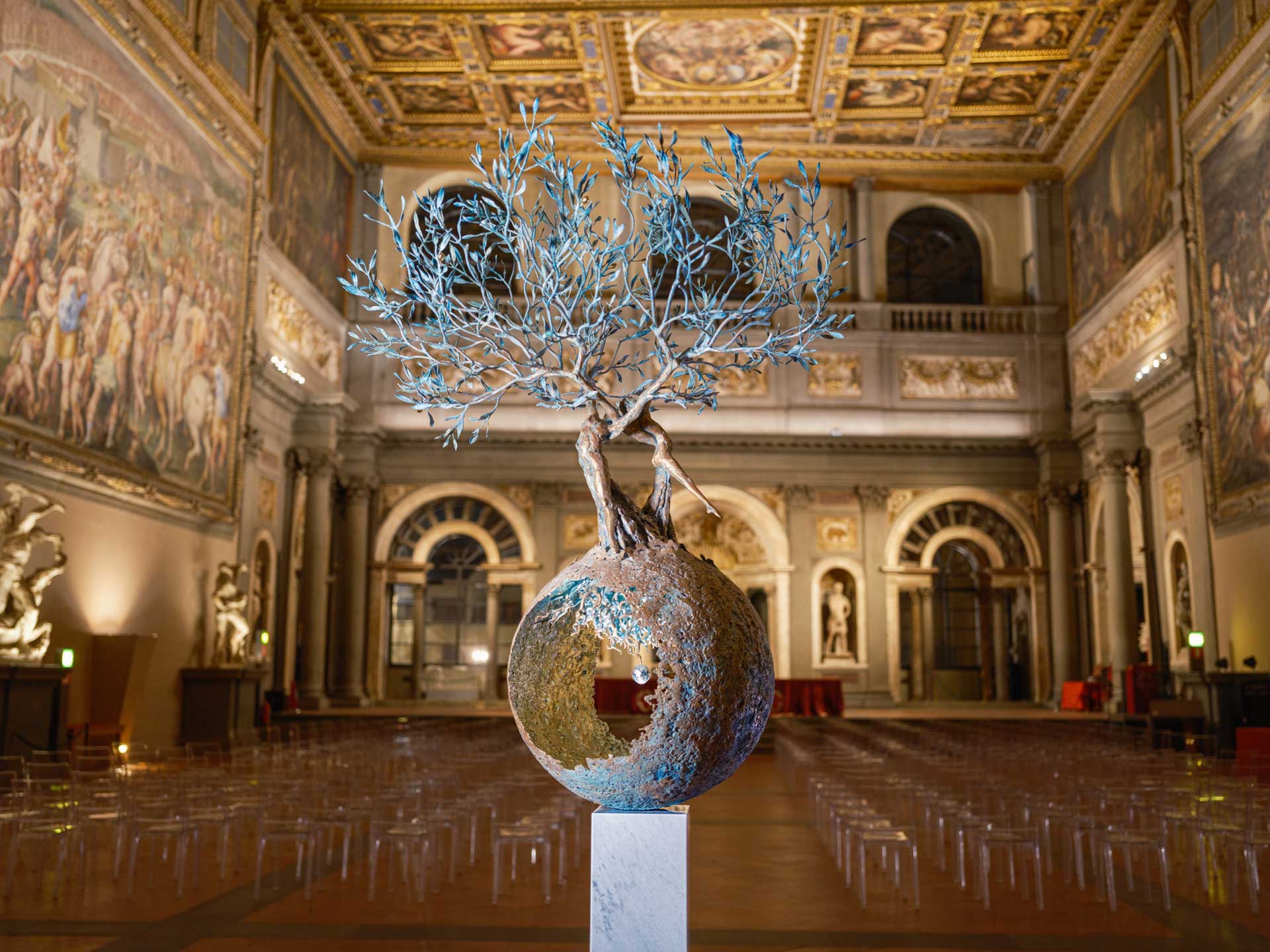 Intricate sculpture of a tree inside a sphere in an ornate hall with detailed murals and decorative ceiling.