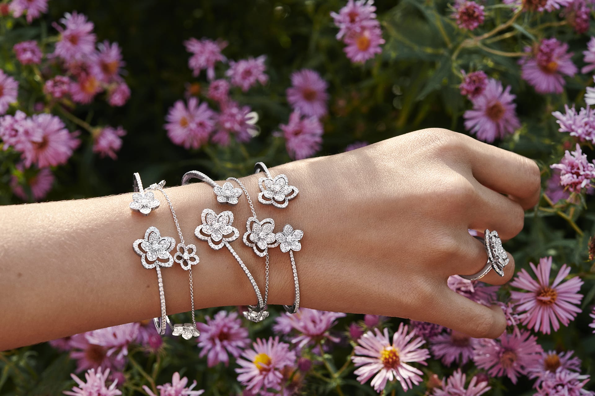 A hand adorned with multiple floral-shaped silver bracelets and a ring, set against a background of blooming pink flowers.