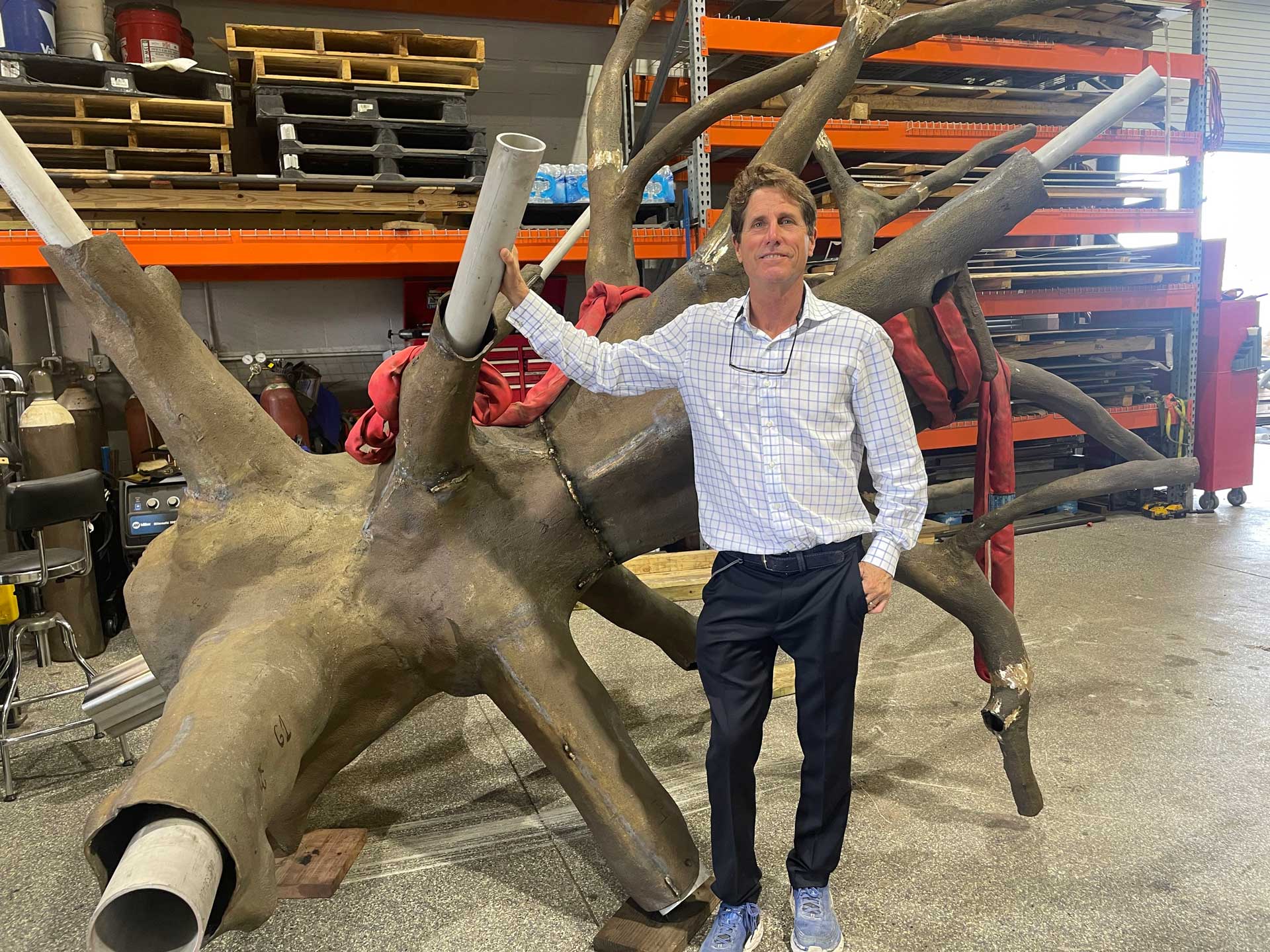 Person standing next to a large, abstract sculpture in a workshop with shelves and tools in the background.