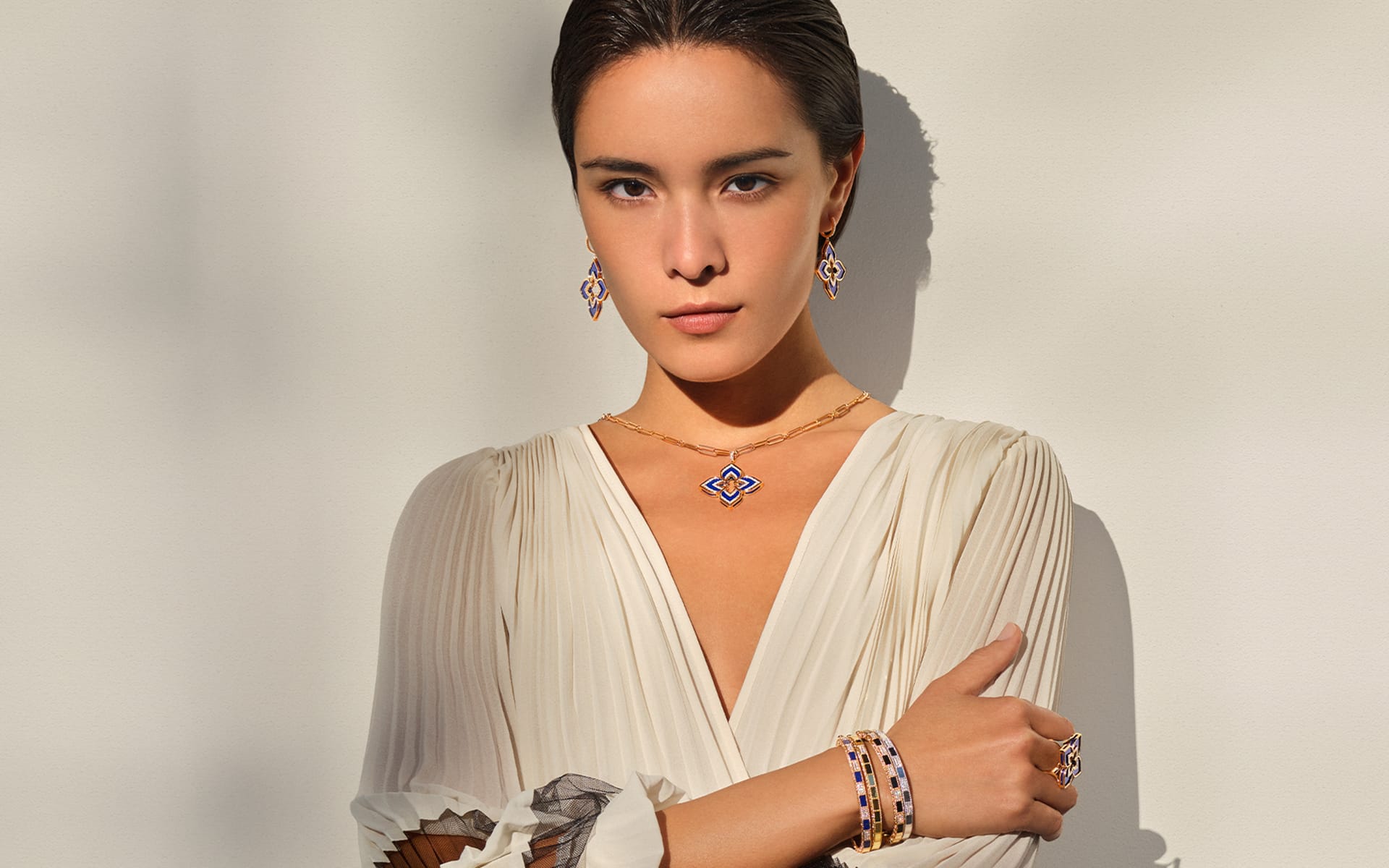 Woman in white dress wearing matching blue and gold jewelry including necklace, earrings, and bracelets, standing against a white wall.