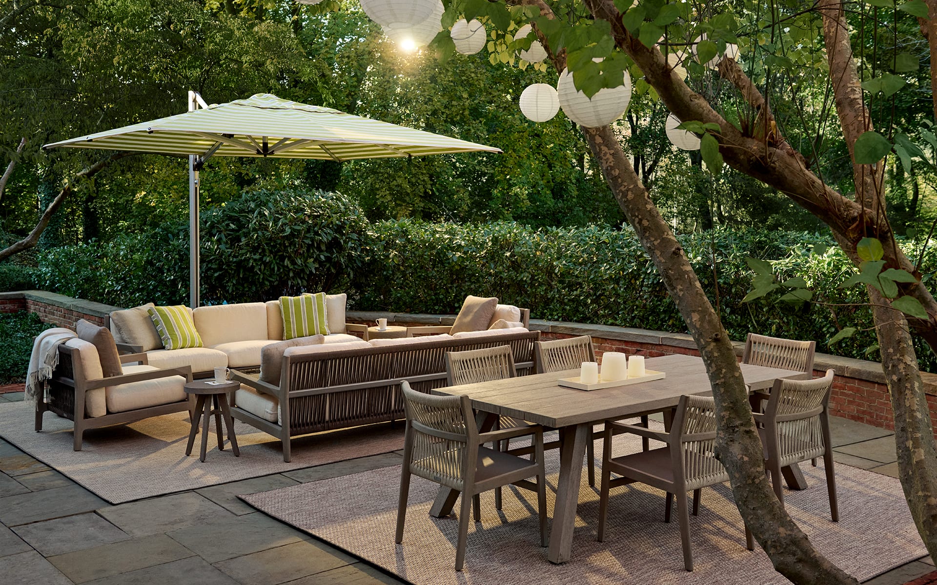 Outdoor patio with dining table, chairs, sofa set, and umbrella surrounded by trees and lanterns.