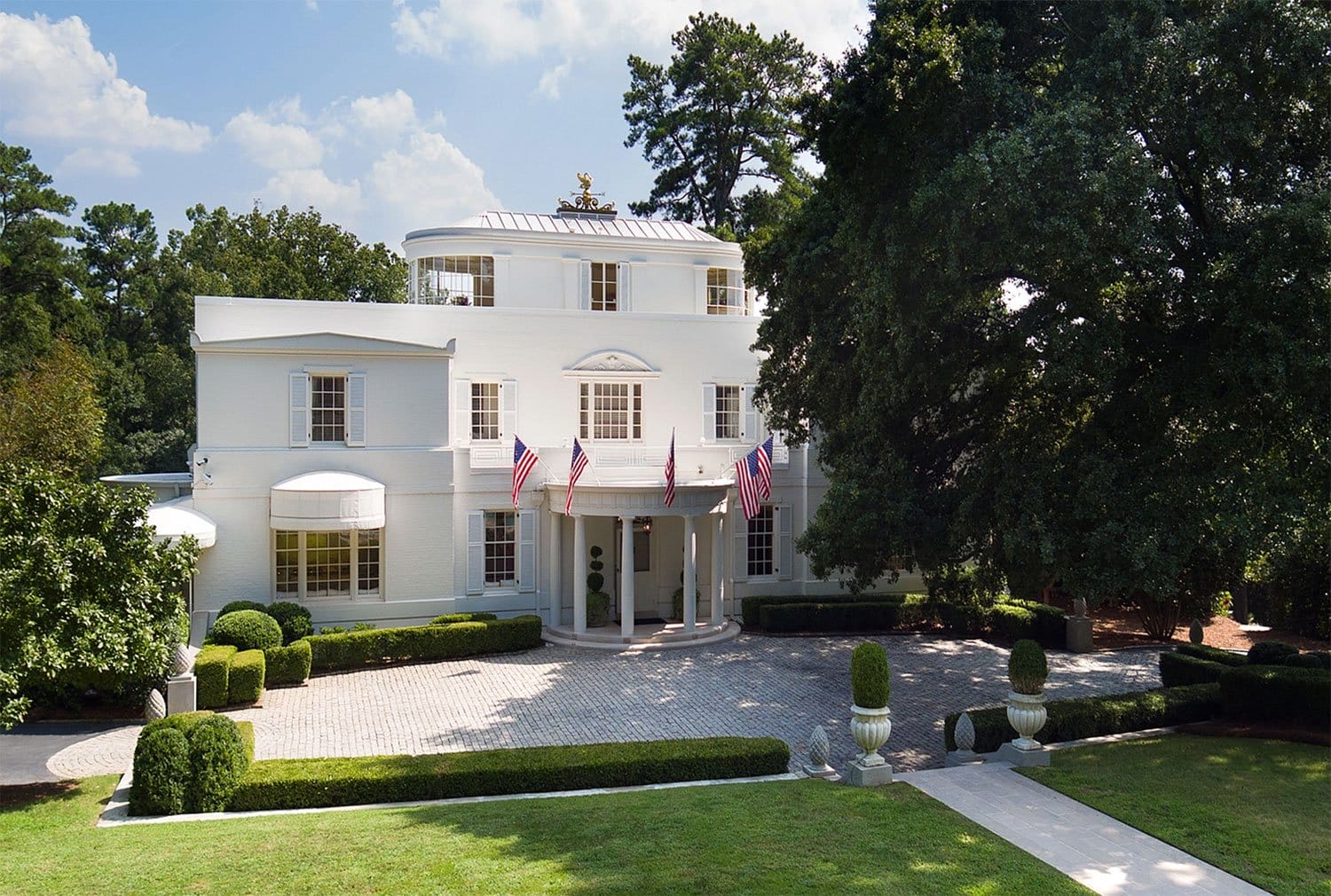 Large white mansion with circular driveway, surrounded by trees and shrubs, adorned with American flags.