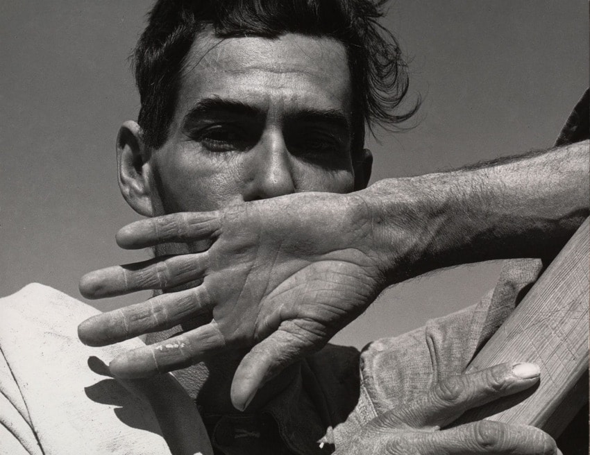 Man shielding face with hand, standing against a clear sky, displaying rugged features and textured skin.