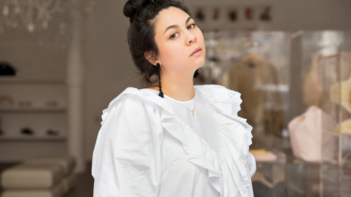 Person in a white ruffled dress poses confidently indoors with blurred clothing displays in the background.