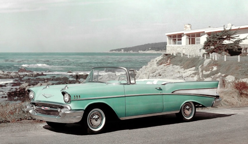 Vintage teal convertible parked by the ocean with a modern house and rocky coastline in the background.