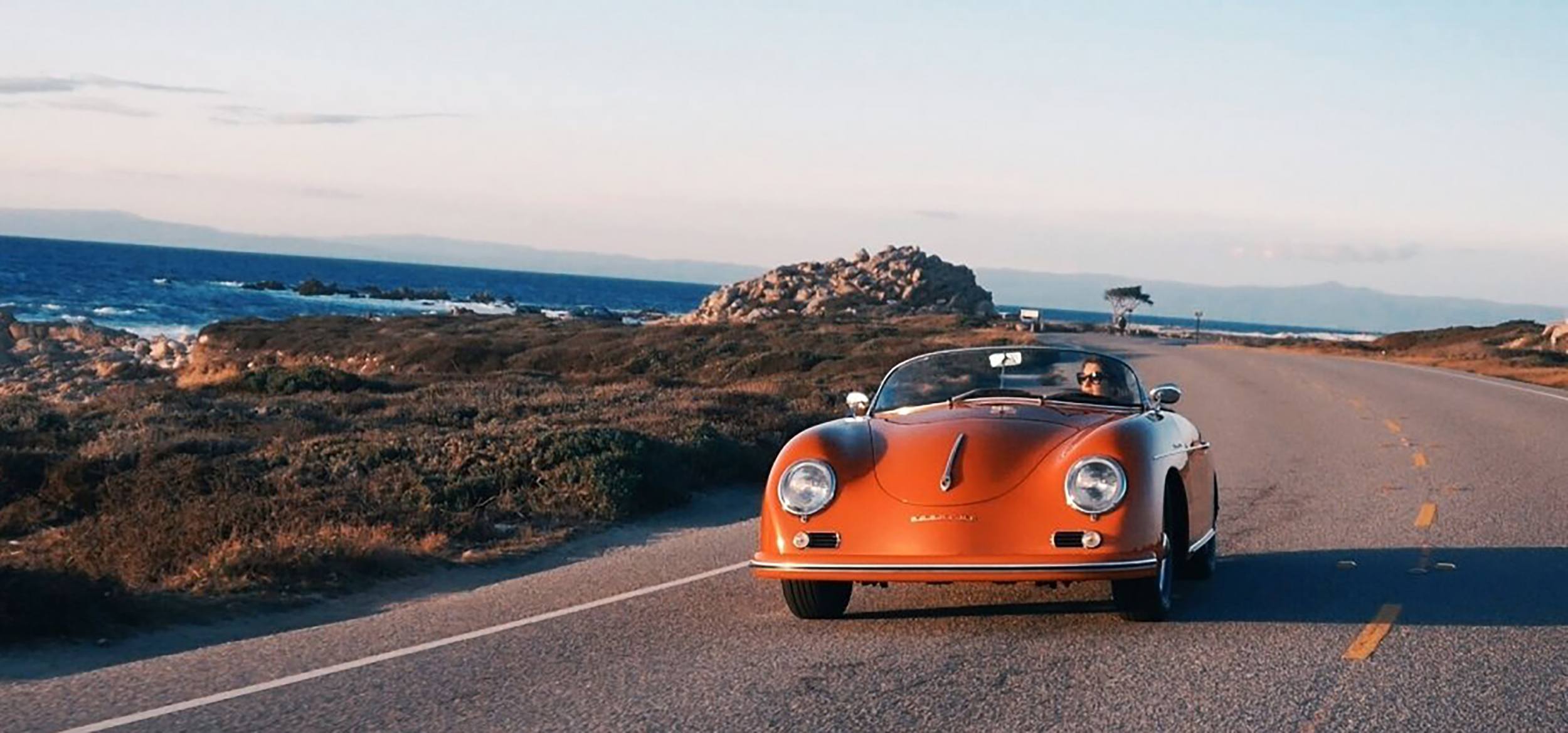 Vintage orange convertible car driving along a scenic coastal road with ocean and rocky landscape in the background.