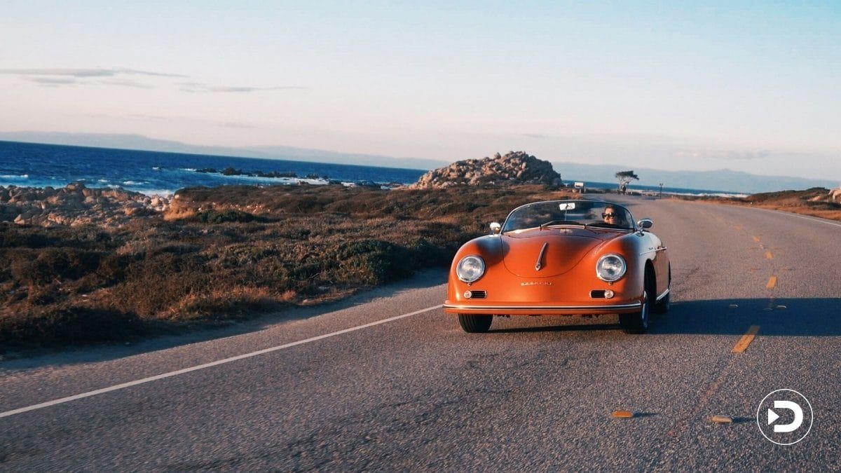 Vintage red convertible driving along a scenic coastal road with ocean view and rocky landscape in the background.