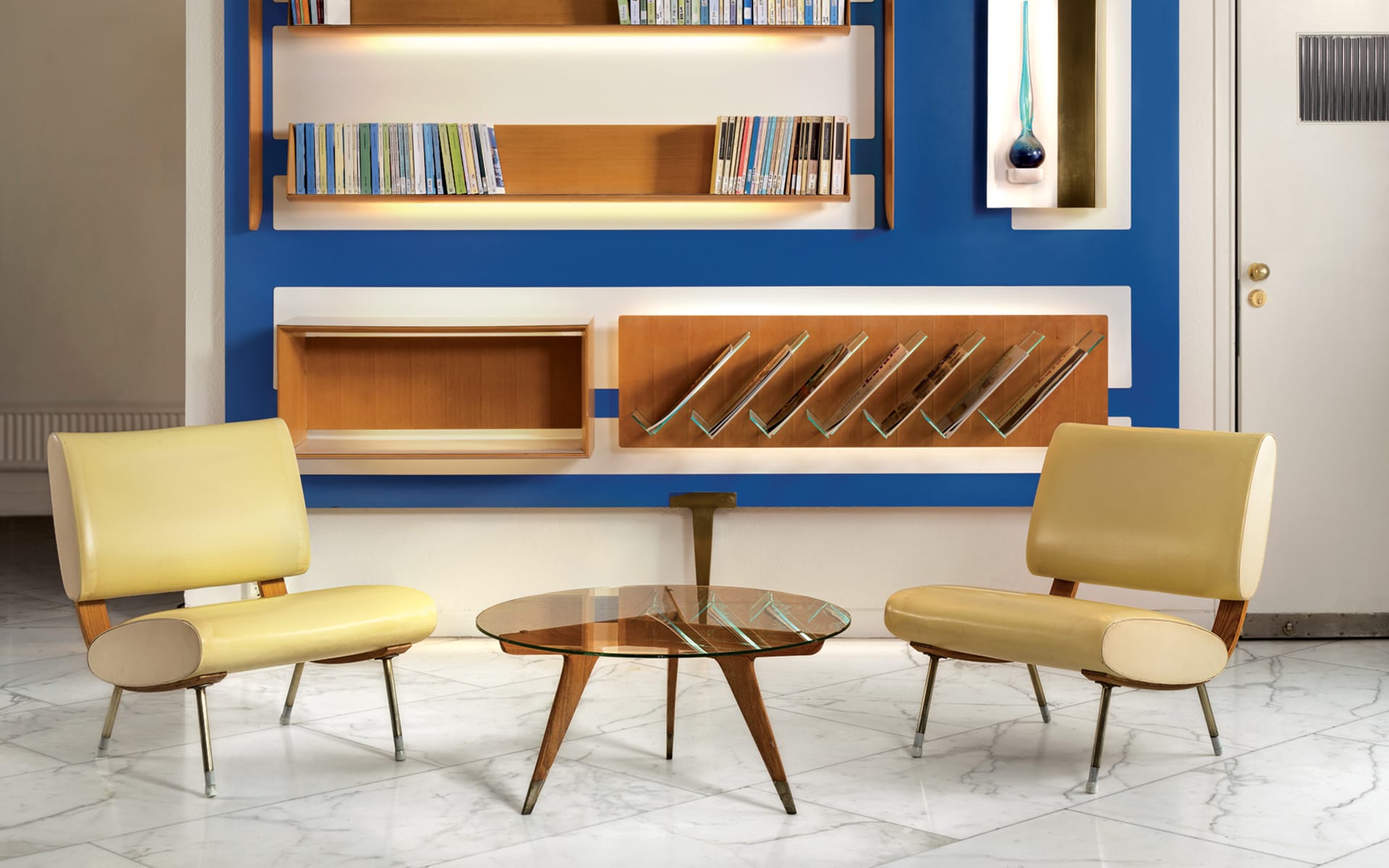 Mid-century modern room with yellow chairs and a glass table, blue wall with wooden shelves and books, marble floor.
