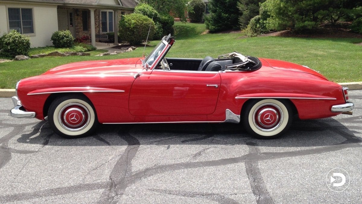 Red vintage convertible car parked on a suburban street with green lawns and houses in the background.