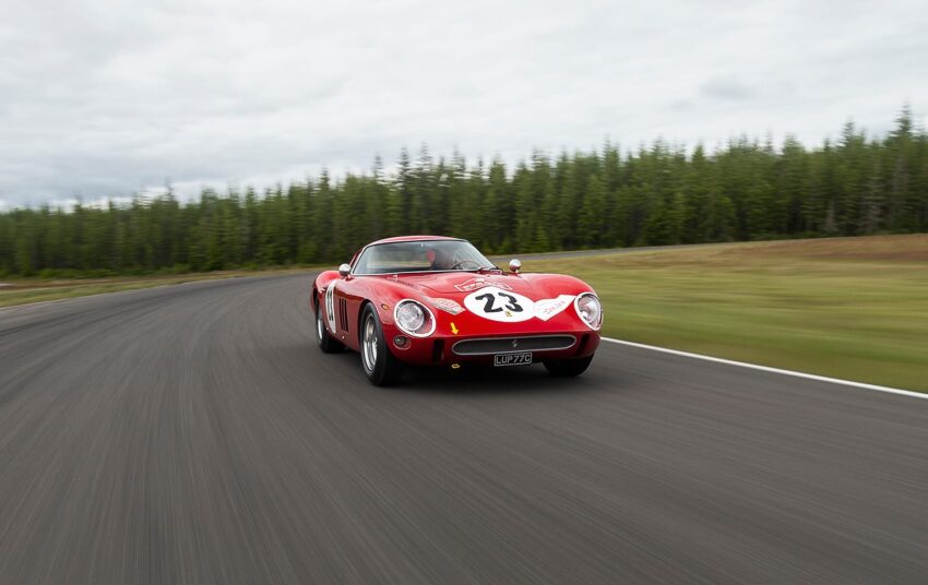 Red vintage sports car with number 23 racing on a track surrounded by trees under a cloudy sky.