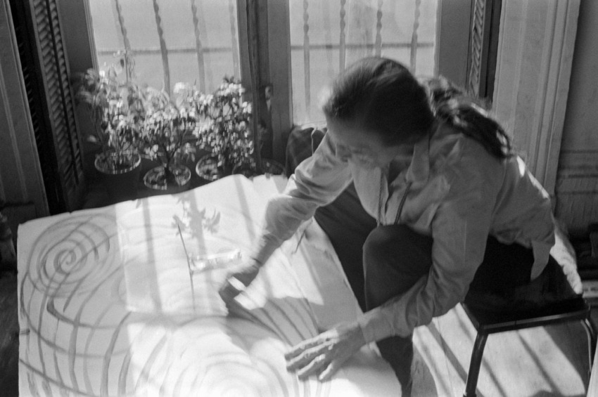 Artist working on abstract circular design by a window with potted plants. Black and white photograph capturing creativity.