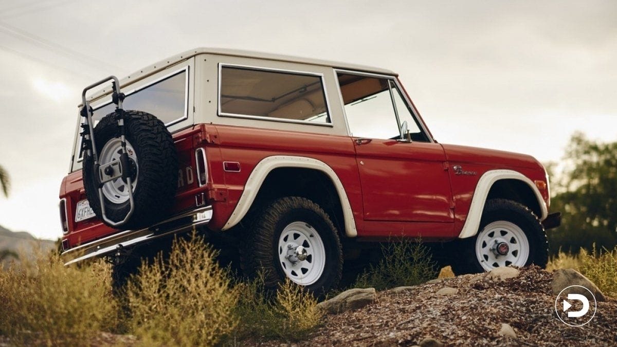 Vintage red off-road vehicle on rocky terrain, rear view showing spare tire and rugged design, surrounded by shrubbery.