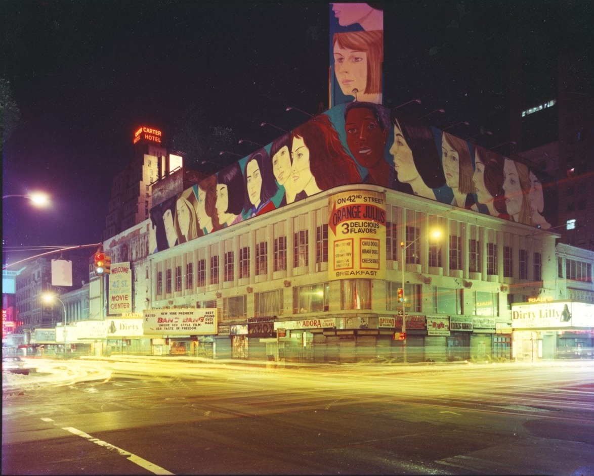 Colorful mural on an urban building at night, showcasing diverse faces, with bright city lights and traffic in the foreground.