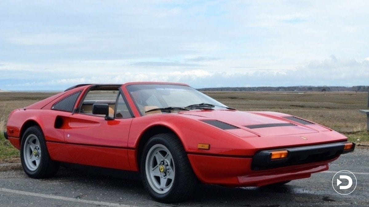 Red classic sports car parked on a scenic countryside road with a partly cloudy sky in the background.