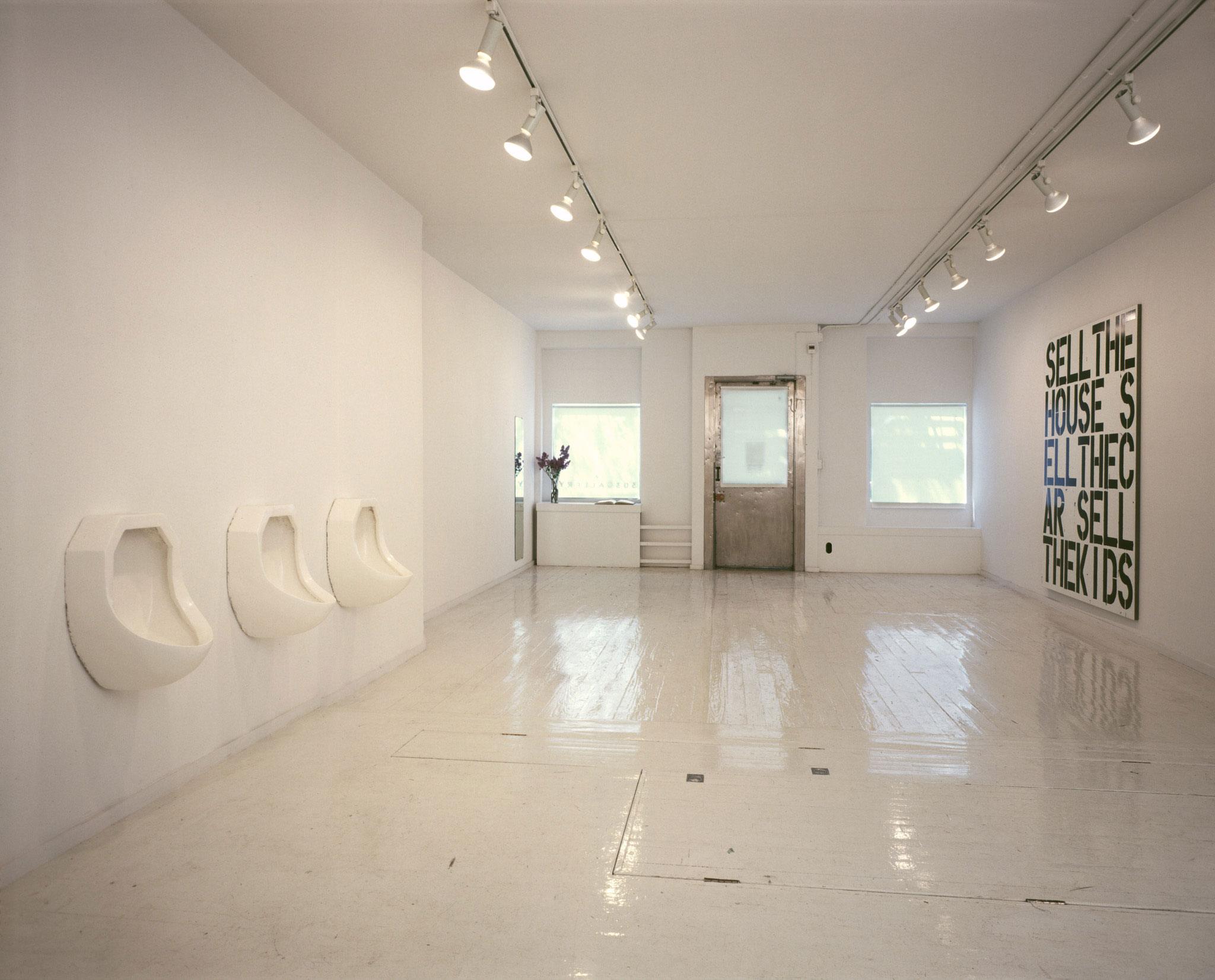 Minimalist gallery with white walls, floor, urinals as art, ceiling lights, and a text-based artwork on the right wall.