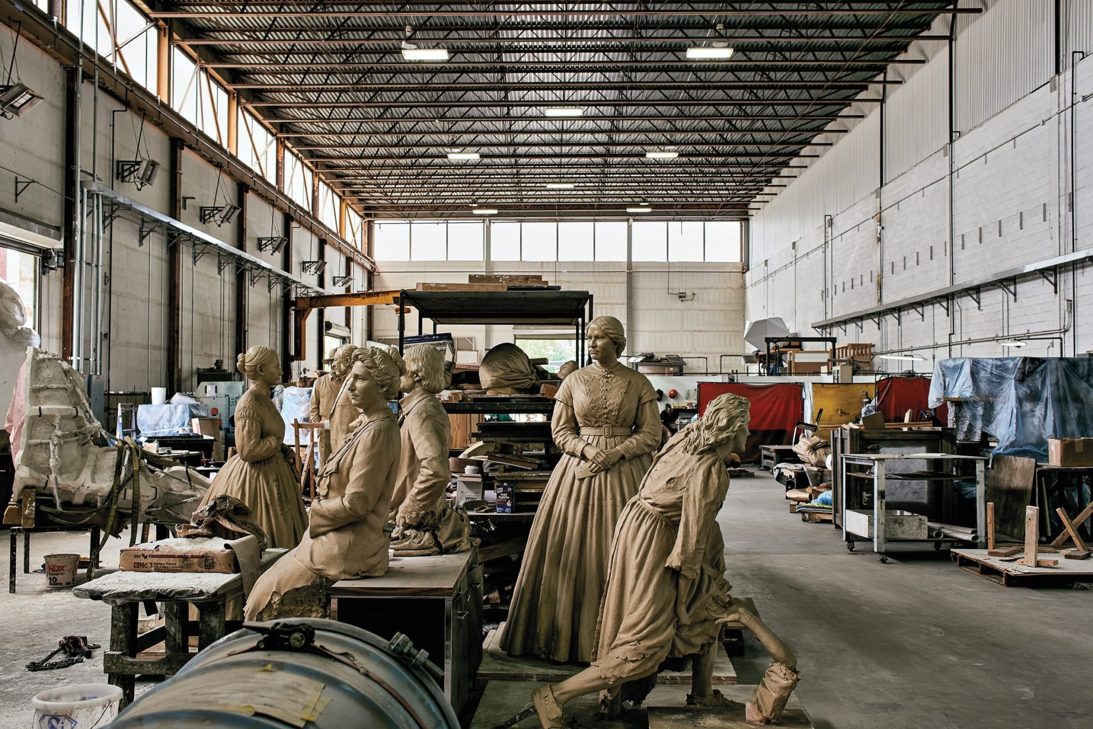 Large warehouse with several unfinished statues of people in historical clothing, surrounded by art supplies and equipment.