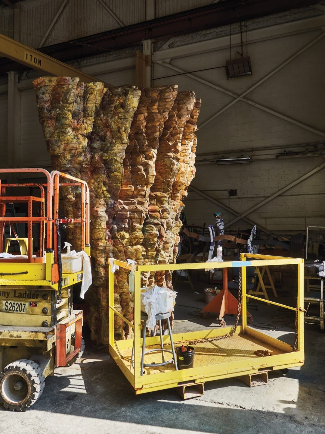 Large abstract sculpture surrounded by scaffolding in an industrial workshop with overhead lighting and various tools around.