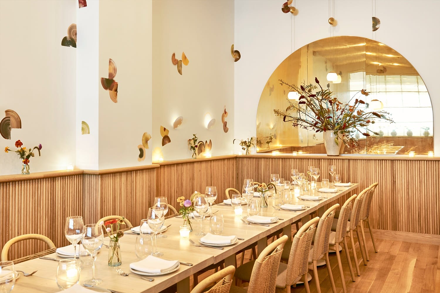 Elegant restaurant interior with a long wooden dining table, cushioned chairs, wall decor, warm lighting, and floral centerpiece.